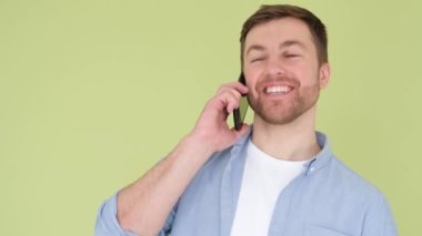 Young handsome man with smartphone in hands, he is talking and laughing isolated on yellow and green studio background. Mobile communication concept