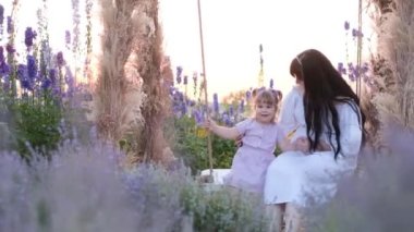 Happy young mother with daughter on a swing in a lavender field. Concept of fun, family, parents, motherhood.