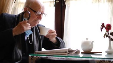 The old grandfather reads the latest news from the newspaper at the table, the pensioner is resting and drinking tea. High quality 4k video