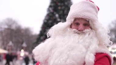 Senior Santa in a red coat shows thumbs up on the background of the Christmas tree as a sign of the thumb up to take a picture on camera. Christmas and New Year concept