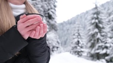 Woman holding a hot cup of tea from a thermos. Rest in the snowy forest. Traveler enjoying hot tea on a frosty winter day. Tourism concept