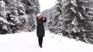 Young woman walking in winter forest on a frosty day. The girl enjoys a walk in the snowy forest and takes pictures of nature on a smartphone. Carefree lady goes and admires the beautiful winter 