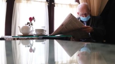 Gri saçlı, gözlüklü ve siyah ceketli yaşlı bir adam pencerenin kenarında bir sandalyede oturuyor, yeni bir gazete okuyor, başka tarafa bakıyor, gazete okuyor. Gerçek zamanlı. 4k video