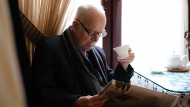 An elderly man is sitting in the kitchen at the table and reading a newspaper. Gray-haired grandfather with glasses sitting on a chair by the window and drinking tea. High quality 4k video