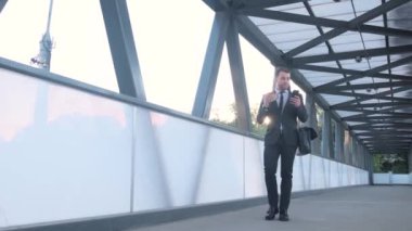 Young businessman in formal clothes looking at the phone while walking down the street and carrying a bag over his shoulder.