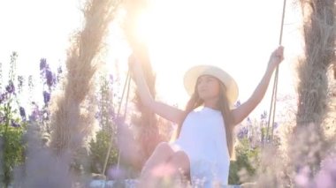 A beautiful young woman is riding on a swing at sunset, she is wearing a beautiful dress and a hat. Summer nature, beautiful purple flowers, sunshine in the frame