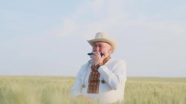 An old Ukrainian grandfather in an embroidered jacket sits on a wheat field and plays the harmonica. Ukrainian traditions