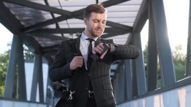 Handsome cheerful man goes to work and looks at the clock. A businessman rushes to the office. Summer sunny morning.