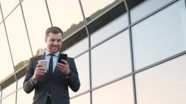 Gülümseyen genç bankacı, elinde bir bardak kahve olan iş adamı bir gökdelenin arkasında duruyor ve telefona bakıyor. İş adamı. Mutlu ve başarılı bir iş adamı.