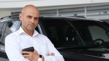 Beyaz gömlekli kel, orta yaşlı bir adam bir araba galerisinde arabanın arka planında duruyor. Araba satıcısı. Satış Yöneticisi.