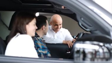 Mutlu genç bir aile, bir araba galerisinde yeni bir arabanın içinde oturuyor ve bir satıcıyla konuşuyor. Araba testi. Araba almak..