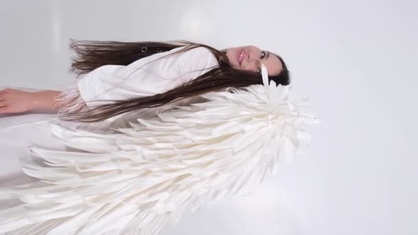 白い背景に白い翼を持つ垂直ビデオ 美しいブルネットの女性 天使の衣装を着た女の子 高品質4Kビデオ — ストック動画