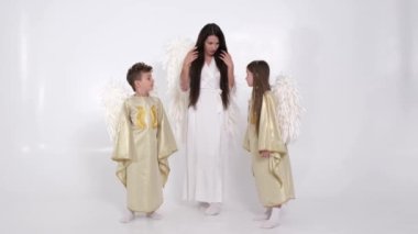 Çocuklu bir anne, melek kostümü giymiş bir kız ve oğul stüdyoda beyaz bir arka planda dans ediyorlar..