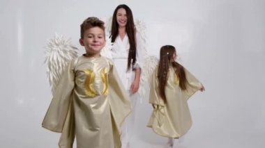 Çocuklu bir anne, melek kostümü giymiş bir kız ve oğul stüdyoda beyaz bir arka planda dans ediyorlar..