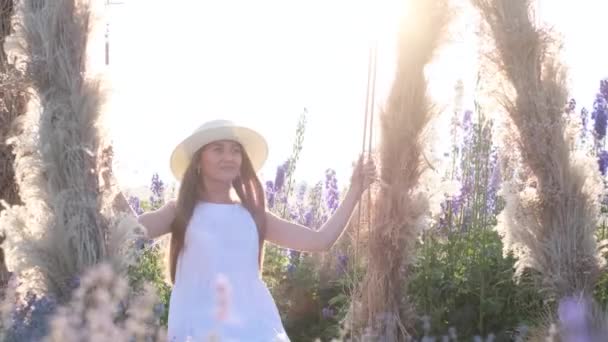 一个穿着白衣的金发姑娘在野花中间荡秋千 在阳光的背景下 在野外拍照 享受大自然带来的快乐 — 图库视频影像