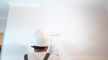 Beyaz kasklı bir inşaat işçisi duvarı beyaza boyuyor. Binanın boyanması ve onarımı. Dairenin tamiri tamamlandı.