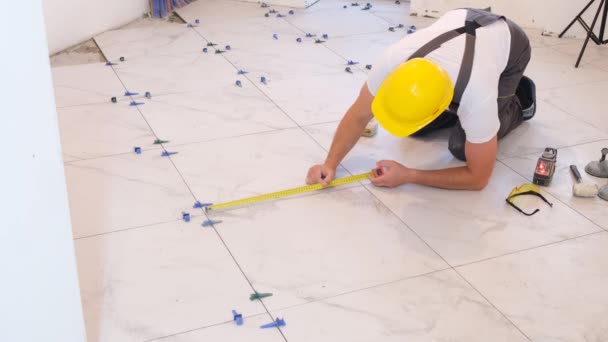 一个穿着工作服的工匠在地板上装瓷砖 在房子里修理 修理公寓 在地板上安装瓷砖 测量和切割零件 — 图库视频影像