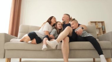 Genç bir aile evdeki kanepede eğleniyor, genç ebeveynler ve çocuklar gülüyor, oynuyor, evde sarılıyor, dinleniyor..