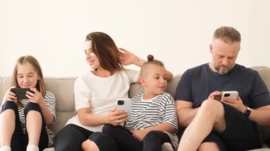 Mutlu Avrupalı aileler, ebeveynler ve küçük çocuklar akıllı telefon cihazlarıyla gülüyorlar, evdeki koltukta birlikte dinleniyorlar, anaokulu çocukları akıllı telefon ekranlarına bakıyorlar, mobil uygulama kullanıyorlar.