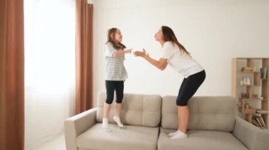 Anne ve kızı kanepede birlikte dans ediyorlar, mutlu bir şekilde zıplıyorlar. Çocuklar ve ebeveynler için neşeli duygular. Mutlu bir aile.
