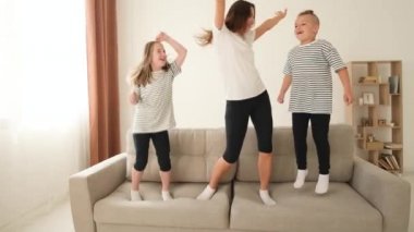 Okul çağındaki anne, kız ve oğlan kanepede dans ediyor, mutlu bir şekilde zıplıyorlar. Aile yatak odasında pijama giyiyor. Çocuk ve ebeveynlerde neşeli duygular.