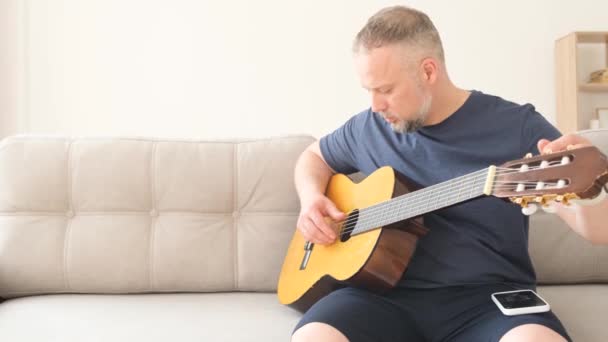 Männlicher Musiker Stimmt Akustikgitarre Während Auf Der Couch Sitzt Entspannt — Stockvideo