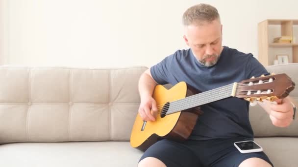 ソファに座っている間 男はアコースティックギターをチューニングします ソファの上の家で休んでいると 夫は妻のために歌う準備をしている 幸せな家族 — ストック動画