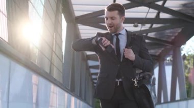 İş merkezinin yakınında genç bir işadamı kol saatine bakıyor. Adam yürür ve gülümser. İş konsepti