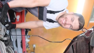 Dikey video. Bir arabayı tamir ederken gülümseyen Avrupa görünümlü çok mutlu bir işçi. İşten neşe. Genç bir adamın olumlu duyguları.