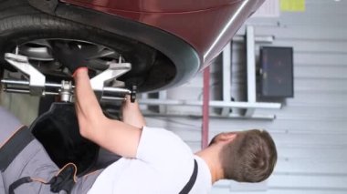 Dikey video. Gri üniformalı genç bir araba tamircisi bir araba tekerleğini söküyor. Atölyede çalış, garajda. Tekerlek değişimi. Otomobil lastikleri