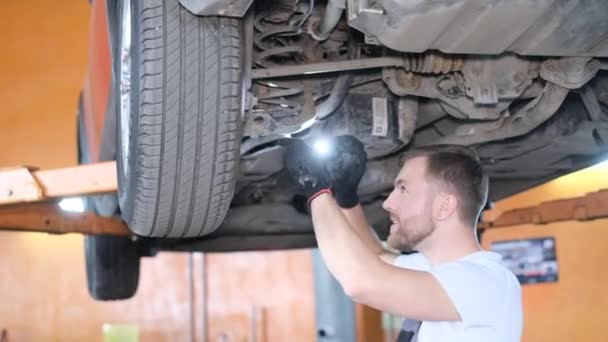 懐中電灯付きの若いアメリカ人の自動車整備士は リフター付きブレーキドラムを検査しています ガレージで車のタイムリーな修理 — ストック動画