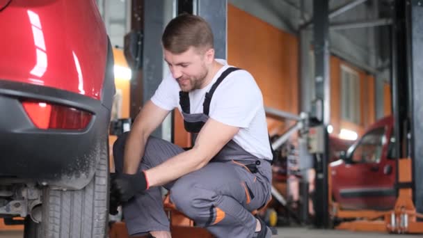 一位身穿灰色制服 面带微笑的年轻汽车修理工正在转动汽车车轮 在车间 车库工作 专业汽车修理 — 图库视频影像
