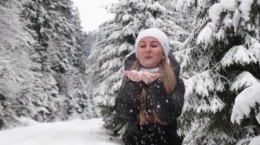 Kış parkındaki bir kız yeşil Noel ağaçlarının arka planında kar taneleri üflüyor, seçici bir odak noktası. Süper yavaş çekim 240 fps