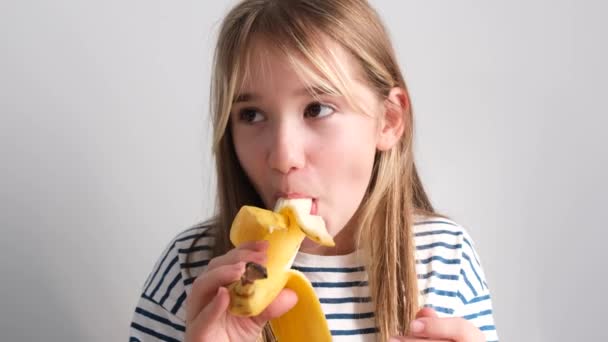 幸せな女の子は黄色のバナナを食べている 7歳の少女は白地にバナナを食べる 4Kビデオ — ストック動画