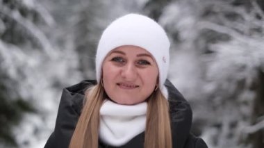 Sıcak beyaz şapkalı genç bir kadının, kış ormanı arka planında, açık havada kameraya bakarken çekilmiş portresini yakından çek. Karlı şehir parkında poz veren mutlu kadın.