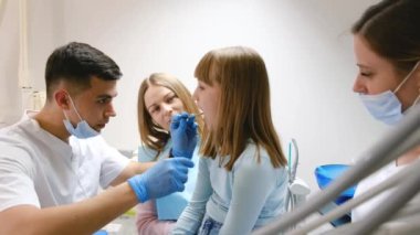 Bir dişçi ve asistanları kızıl saçlı genç bir kızın ağız boşluğunu inceliyor. Gençlerde diş tedavisi uygulanıyor. Profesyonel diş tedavisi