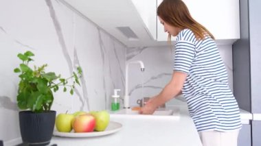 Avrupalı görünümlü genç bir kadın modern beyaz bir mutfakta bulaşıkları yıkıyor. Hafta içi evde.
