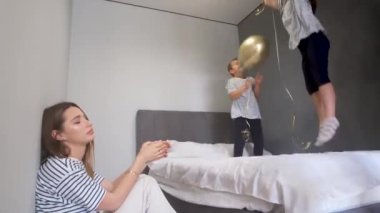 Yorgun anne ve mutlu çocuklar. Bir erkek ve bir kız balonlarla bir yatakta zıplıyorlar. Çocuklar yaramazlık yapar