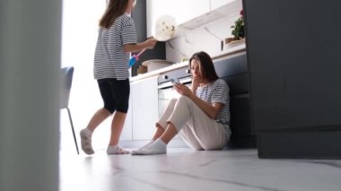 Genç bir kadın modern bir mutfakta yerde otururken telefona bakıyor. Genç anne ve kızı. Balonlu bir çocuk.