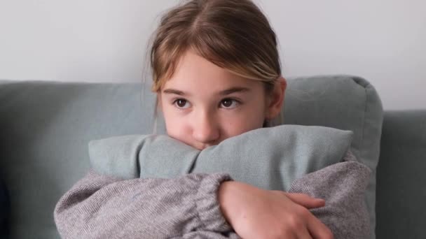 金髪の女の子がテレビを見ている かわいい女の子の肖像画はソファの上に座って 灰色の枕にもたれてまっすぐ前を見る — ストック動画