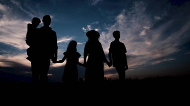 Gün batımının arka planında bir aile yürüyor, el ele tutuşuyor, gün batımının arka planında siluetler var. Mutlu ve güçlü bir aile..