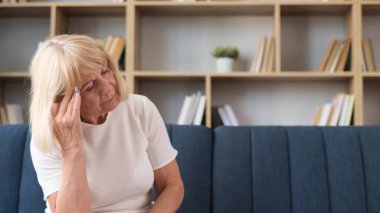 Sad elderly woman, grandmother sitting on sofa in living room and suffering from headache. High blood pressure. Health problems.
