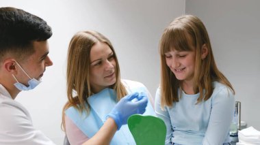 Çocuklarla dişçiye gidiyorum. Küçük bir kız, klinikte dişçi koltuğunda annesiyle birlikte oturuyor. Doktor, bebek dişlerinin incelenmesi için hazırlanıyor..
