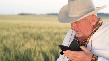 Ukrainian grandfather is talking on a smartphone, holding it in his hand. Mobile communication in the middle of a field in the countryside