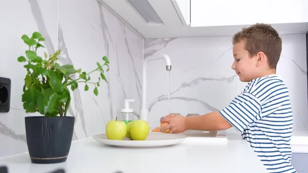 A little boy washes fruit under tap water. Child hands holding tasty apple under running water in kitchen sink.