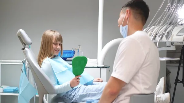 坐在医疗椅上的快乐小女孩照着镜子 一个戴着防护面具 身穿制服的年轻医生看着孩子 — 图库照片