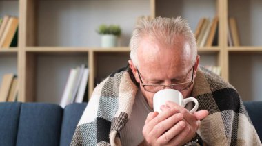 A gray-haired grandfather is sitting on the sofa, covered with a gray blanket and drinking warm tea. Grandfather has the flu. Have a nice time at home.
