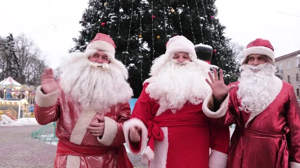 Three Santa Clauses on the background of a Christmas tree in the city center. Group of Santa Clauses waving at the camera. Christmas and New Year concept