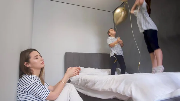 Tired mother and happy children. A boy and a girl jump on a bed with balloons. Childrens mischief