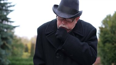 Parktaki olgun adam göğsünü tutar ve yakalar. Yaşlı adam parkta öksürüyor.